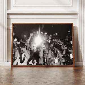 May include: A framed black and white photograph featuring a crowd of photographers with cameras and flashes. The image is in a brown wooden frame, set against a white wall and a wooden floor.