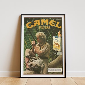 May include: Framed vintage Camel Filters advertisement. The poster features a man smoking a cigarette, with a pack of Camel cigarettes and the brand's logo. The background includes lush green foliage.