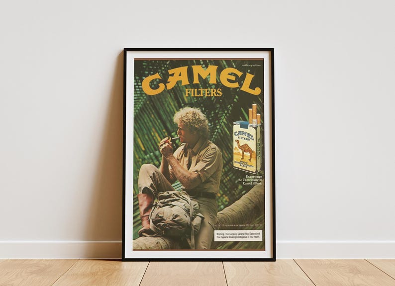 May include: Framed vintage Camel Filters advertisement. The poster features a man smoking a cigarette, with a pack of Camel cigarettes and the brand's logo. The background includes lush green foliage.