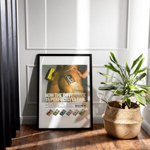May include: Framed vintage Sony advertisement poster. The poster features a retro image with the text "NOW THE DIFFERENCE IS PERFECTLY CLEAR." Below the main image are several colorful cassette tape designs. A potted plant in a woven basket sits nearby.