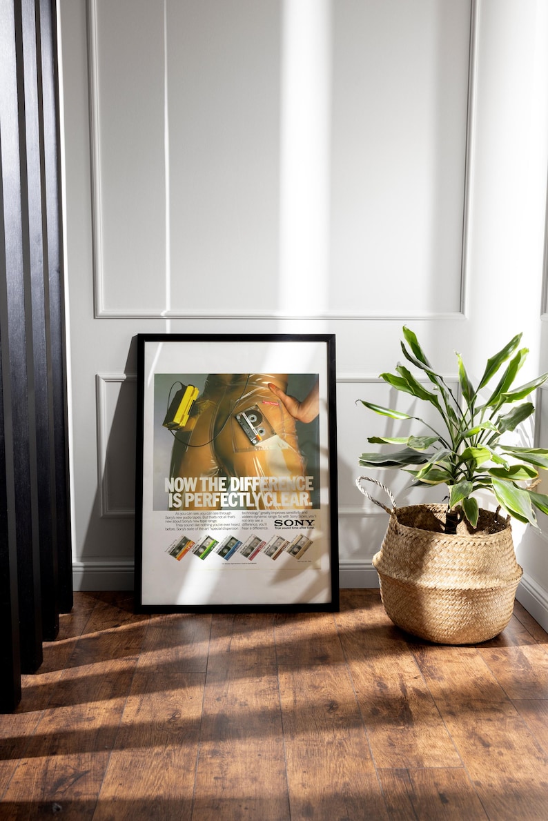 May include: Framed vintage Sony advertisement poster. The poster features a retro image with the text "NOW THE DIFFERENCE IS PERFECTLY CLEAR." Below the main image are several colorful cassette tape designs. A potted plant in a woven basket sits nearby.