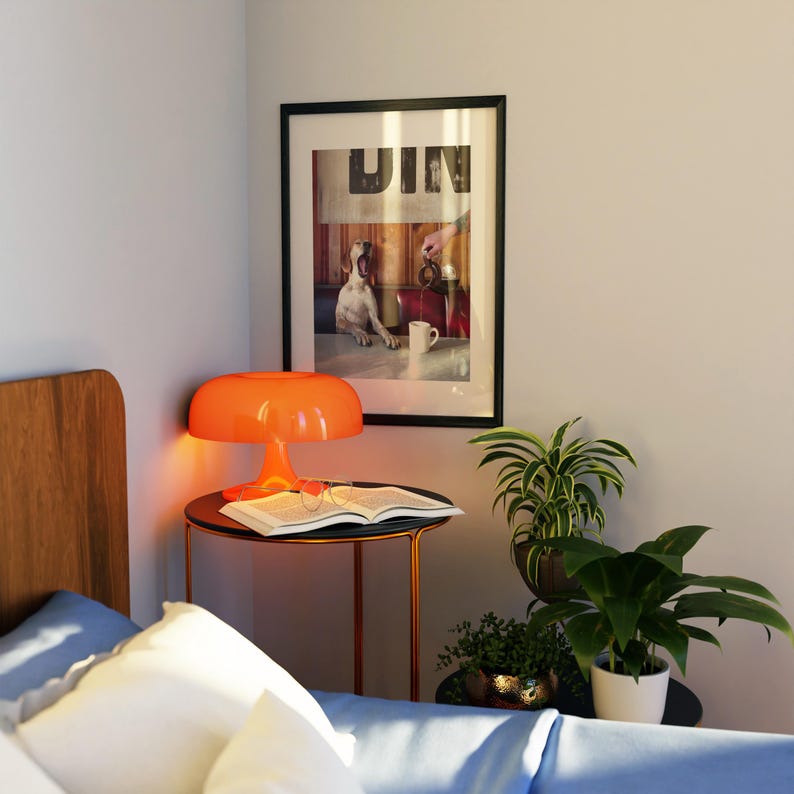 May include: An orange mushroom-shaped lamp sits on a black side table next to a bed. A framed picture with the word "DIN" is on the wall. A book and glasses are on the table. Potted plants are on the table and floor.