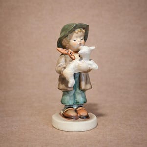 May include: A ceramic figurine of a child in a green hat and coat holding a white lamb. The figurine has a brown and tan color scheme, with the child's face and hands visible. The figurine is standing on a round, off-white base.