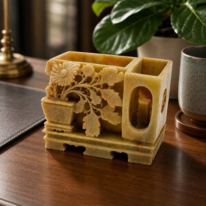May include: A rectangular beige stone desk organizer featuring intricate relief carvings of a potted chrysanthemum flower on the front panel. The piece includes two distinct compartments for storage and rests on a carved base with cutout details.