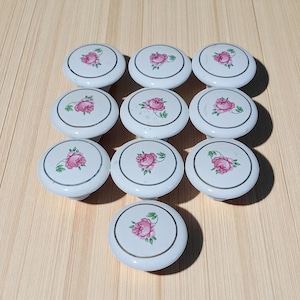 May include: A collection of ten white ceramic cabinet knobs. Each knob features a pink rose design in the center, surrounded by a silver-colored ring. The knobs are arranged on a light-colored wooden surface.