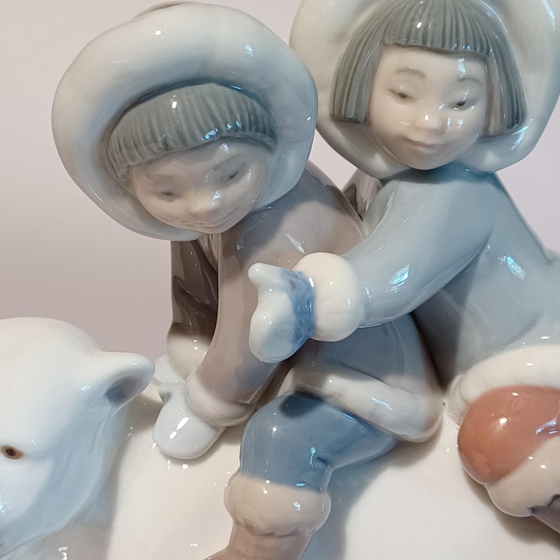 Pu&ograve; includere: Una statuina di porcellana raffigurante due bambini in abiti invernali, con cappotti e cappelli azzurri, e un orso polare. I bambini interagiscono con l'orso polare. La statuina ha una finitura lucida ed &egrave; ambientata su sfondo bianco.