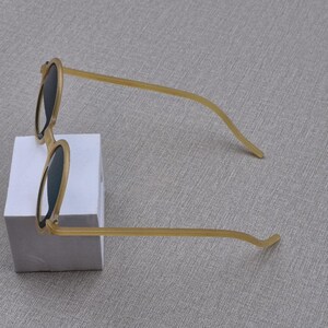 May include: Round sunglasses with a light brown frame and dark lenses. The sunglasses are on a white block against a grey background. The arms are thin and curve. The frame is a light tan colour.