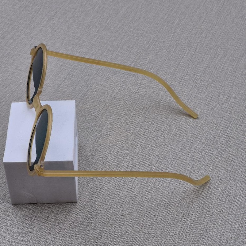 May include: Round sunglasses with a light brown frame and dark lenses. The sunglasses are on a white block against a grey background. The arms are thin and curve. The frame is a light tan colour.