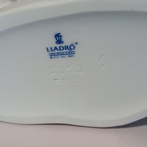 Pu&ograve; includere: Base di statuetta in porcellana bianca con testo e logo blu. Il testo include "LLADR&Oacute; HAND MADE IN SPAIN &copy; DAISA 1985" e i numeri "5353" e "7-10 D". Il logo &egrave; un emblema blu.