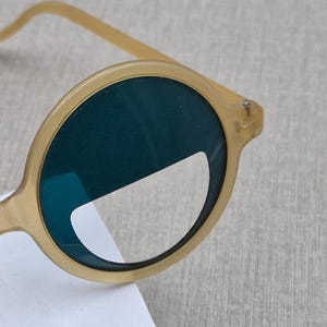 May include: Close-up of round sunglasses with a light tan frame and dark green lenses. The sunglasses have a unique, modern design. The lenses are partially covered by a white sticker.