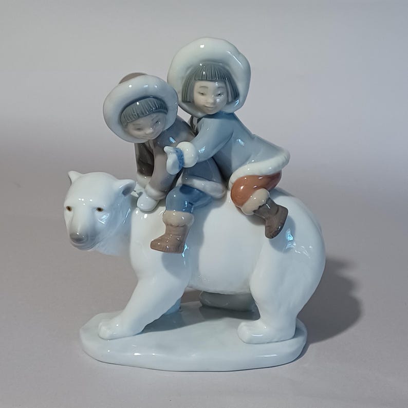 Pu&ograve; includere: Statuina in porcellana raffigurante due bambini che cavalcano un orso polare. I bambini indossano cappotti invernali blu e marroni con finiture in pelliccia bianca e cappelli. L'orso polare &egrave; bianco con un naso grigio chiaro.