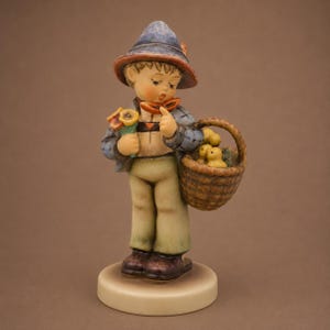 May include: A hand-painted ceramic figurine of a child wearing a blue hat and jacket, tan trousers, and brown shoes. The child holds a small bouquet of yellow flowers in one hand and carries a woven brown basket containing three yellow chicks.