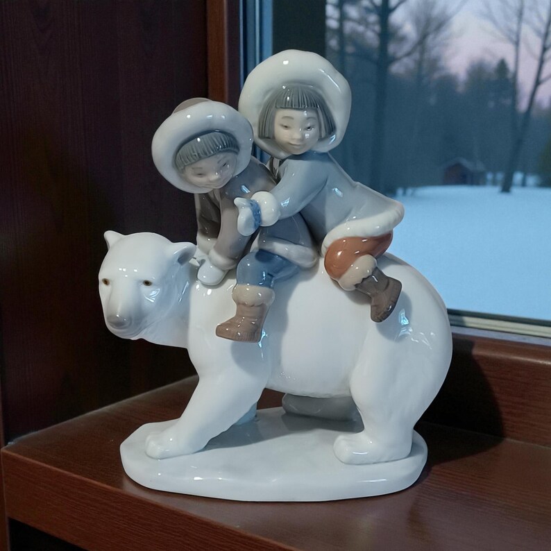 Pu&ograve; includere: Statuina in ceramica bianca raffigurante due bambini che cavalcano un orso polare. I bambini indossano cappotti e cappelli invernali nei toni del grigio, blu e marrone. L'orso polare &egrave; bianco. La statuina &egrave; posta su una base bianca. Misure in centimetri.