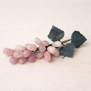May include: A decorative cluster of pink and white gemstone grapes with green leaves and a stem. The grapes vary in shades of pink, from light to deep rose. The leaves are a dark green color.