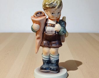 May include: A ceramic figurine of a young boy in traditional German attire, holding a large cone-shaped object and a small satchel. The boy wears a green jacket, brown shorts, and black boots. The figurine is on a round, off-white base.