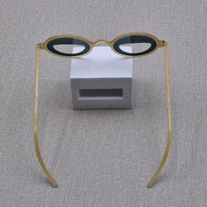 May include: Oval sunglasses with a gold frame and dark lenses. The glasses are displayed on a white rectangular block against a grey background. The thin arms extend outwards.