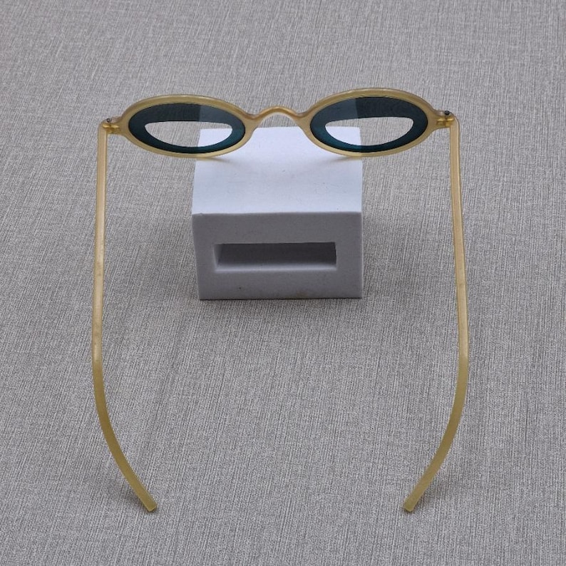 May include: Oval sunglasses with a gold frame and dark lenses. The glasses are displayed on a white rectangular block against a grey background. The thin arms extend outwards.