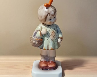May include: A ceramic figurine of a young child holding a basket. The child wears a light green dress with a diamond pattern and brown shoes. An orange accent sits atop the child's head. The figurine is on a white base.