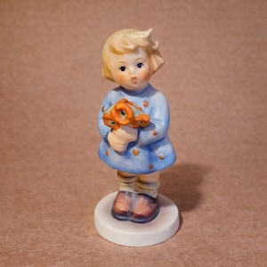 May include: A ceramic figurine of a young girl in a blue dress holding a bouquet of orange and blue flowers. The figurine has blonde hair, rosy cheeks, and is standing on a white circular base. The dress has small blue dots.
