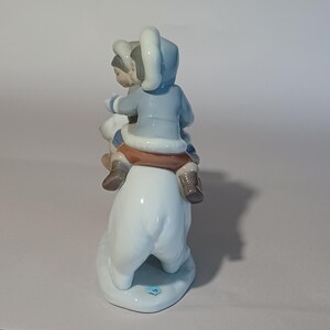 Pu&ograve; includere: Statuina in porcellana raffigurante due bambini che cavalcano un orso polare. I bambini indossano cappotti con cappuccio azzurri, pantaloni marroni e stivali marroni. L'orso polare &egrave; bianco. La statuina &egrave; su una base bianca.