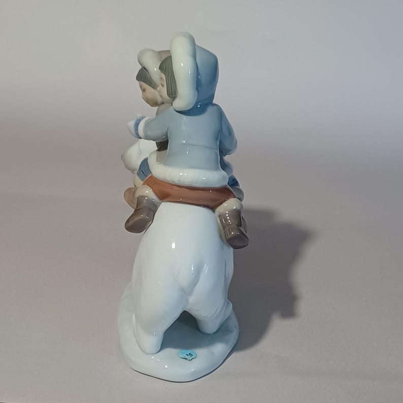 Pu&ograve; includere: Statuina in porcellana raffigurante due bambini che cavalcano un orso polare. I bambini indossano cappotti con cappuccio azzurri, pantaloni marroni e stivali marroni. L'orso polare &egrave; bianco. La statuina &egrave; su una base bianca.