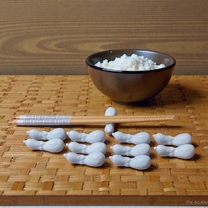 May include: A brown bowl filled with white rice sits on a bamboo mat. Chopsticks rest on a white ceramic chopstick rest. Several white ceramic chopstick rests are arranged in front of the chopsticks. The background is a wooden surface.