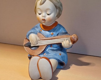 May include: A ceramic figurine of a seated child playing a mandolin. The child wears a blue outfit with orange trim and has closed eyes. The mandolin is brown with black strings. The figurine is on a light-coloured surface.
