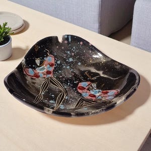 May include: A black decorative dish with a unique shape and painted figures. The dish features a dark background with colorful splatters and two figures in pink, blue, and black. A stylish accent piece.