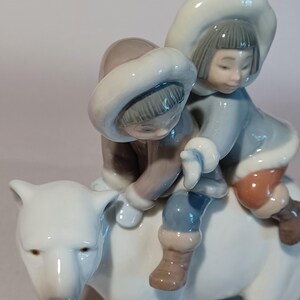 Pu&ograve; includere: Statuina in porcellana raffigurante due bambini che cavalcano un orso polare. I bambini indossano abiti invernali azzurri e marroni con cappucci foderati di pelliccia bianca. L'orso polare &egrave; bianco con un volto dettagliato. La statuina ha una finitura lucida.