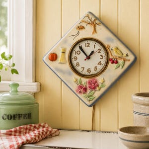 May include: A diamond-shaped wall clock with a cream-colored face featuring black numerals and hands. The clock is decorated with raised relief figures including a pumpkin, a wheat sheaf, a bare tree, a yellow bird on a branch, and pink flowers.