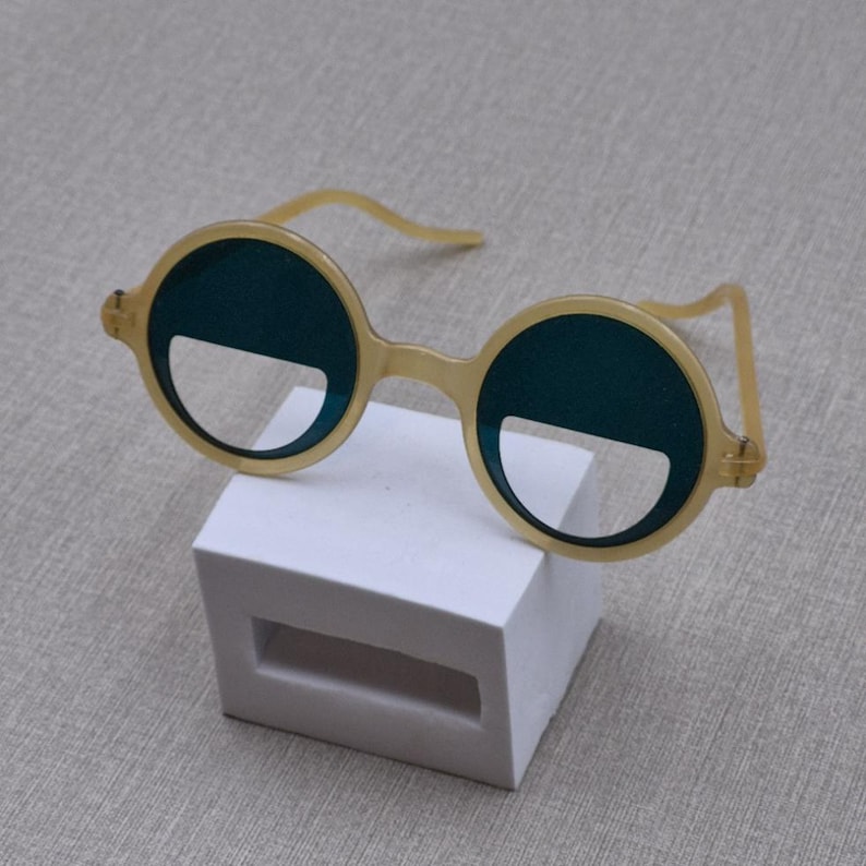May include: Round, yellow-framed sunglasses with dark teal lenses and a distinctive half-circle cutout at the bottom. The glasses are displayed on a white rectangular stand. A fashionable accessory for a retro or modern style.