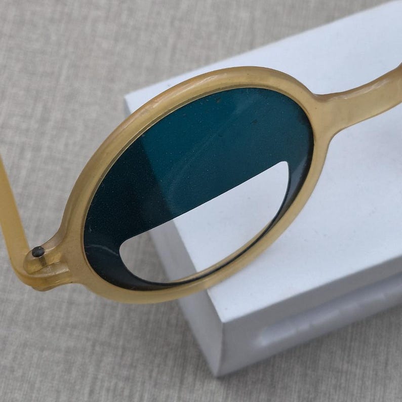 May include: Close-up of a pair of round, light-yellow sunglasses with dark teal lenses. The frame is a solid colour, and the lenses have a unique shape. The sunglasses are resting on a white surface.