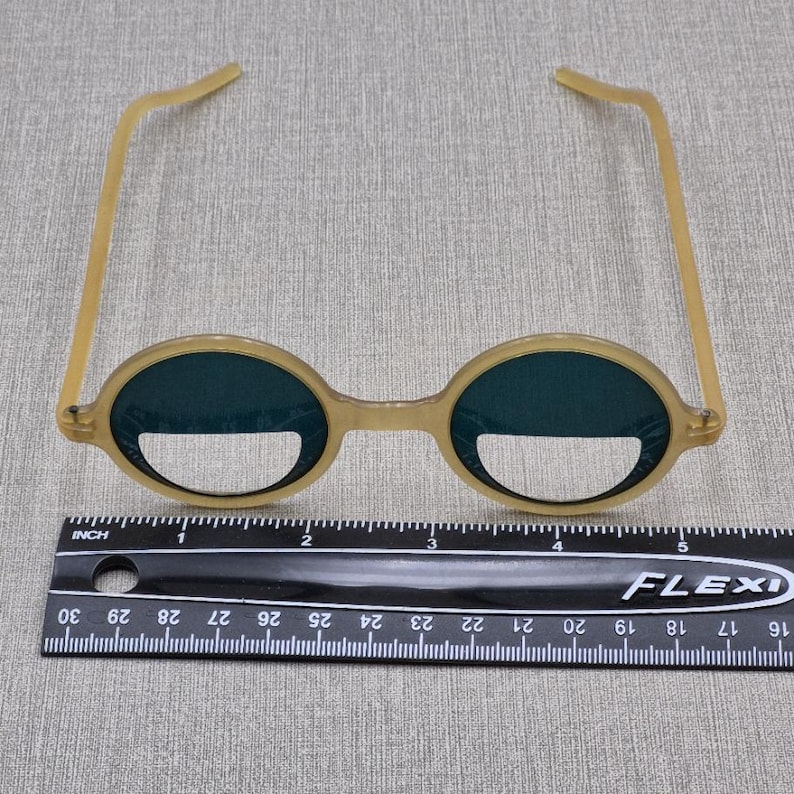 May include: Tan-coloured, round-framed glasses with dark green lenses. The glasses have a bridge and arms, and are placed on a surface with a ruler. The glasses are approximately 13 cm wide.