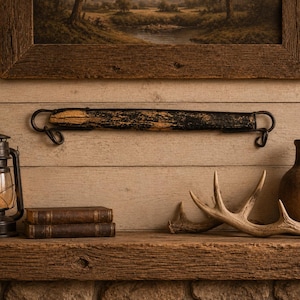 May include: A rustic shelf display featuring a vintage oil lamp, two leather-bound books, a decorative antler, and a brown vase. A distressed wooden beam with metal hooks hangs above the shelf, with a framed landscape painting in the background.