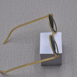 May include: Round sunglasses with a light brown frame and dark lenses. The sunglasses are positioned on a white block against a grey background. The arms of the sunglasses are thin and extend outwards.