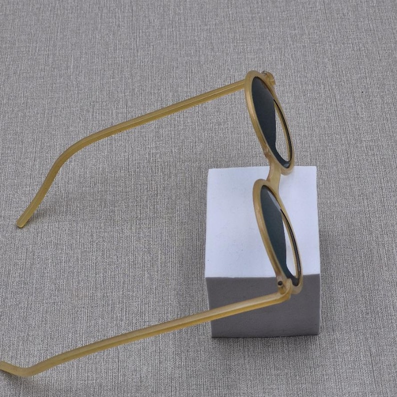 May include: Round sunglasses with a light brown frame and dark lenses. The sunglasses are positioned on a white block against a grey background. The arms of the sunglasses are thin and extend outwards.