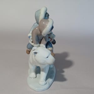 Pu&ograve; includere: Statuina in ceramica raffigurante tre figure su un orso polare. Le figure indossano abiti invernali blu, grigi e marroni. L'orso polare &egrave; bianco, con una base azzurra. Oggetto decorativo.