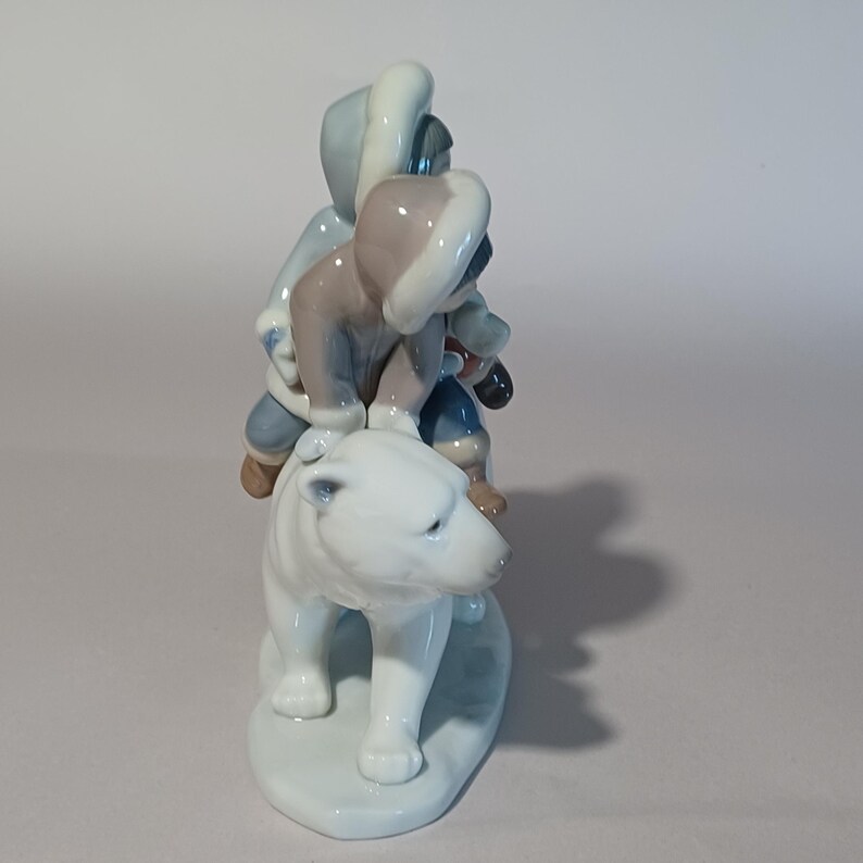 Pu&ograve; includere: Statuina in ceramica raffigurante tre figure su un orso polare. Le figure indossano abiti invernali blu, grigi e marroni. L'orso polare &egrave; bianco, con una base azzurra. Oggetto decorativo.