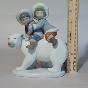 Pu&ograve; includere: Statuina in porcellana raffigurante due bambini a cavallo di un orso polare. I bambini indossano cappotti e cappelli invernali blu e grigi. L'orso polare &egrave; bianco con una base grigia. La statuina &egrave; alta circa 13 cm.