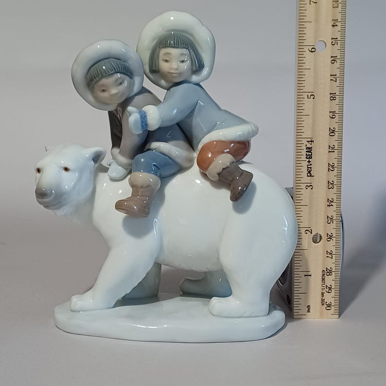 Pu&ograve; includere: Statuina in porcellana raffigurante due bambini a cavallo di un orso polare. I bambini indossano cappotti e cappelli invernali blu e grigi. L'orso polare &egrave; bianco con una base grigia. La statuina &egrave; alta circa 13 cm.