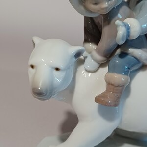 Pu&ograve; includere: Statuina in porcellana raffigurante un bambino che cavalca un orso polare. Il bambino indossa un cappotto grigio con cappuccio, pantaloni blu e stivali marroni. L'orso polare &egrave; bianco con occhi marroni. La statuina ha una finitura lucida.