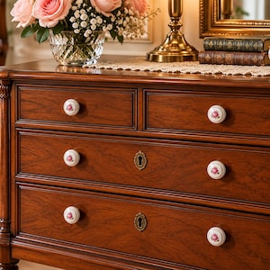 May include: A wooden chest of drawers featuring a rich brown finish, five drawers, and white ceramic knobs decorated with small pink floral patterns. The two lower drawers include brass-toned keyhole escutcheons centered on the drawer fronts.
