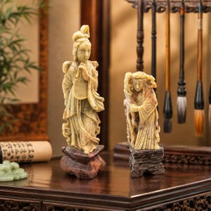 May include: Two carved stone figurines depicting figures in traditional robes, each standing on a dark wooden base. The taller figure holds a fan-like object, while the shorter figure stands beside a textured element. Both are light beige in color.