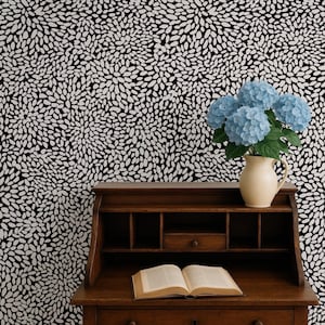 May include: A dark wood roll-top desk with an open book on the surface and a cream-colored pitcher containing blue hydrangea flowers. The background features a wallpaper with a repeating pattern of small white oval shapes on a black field.