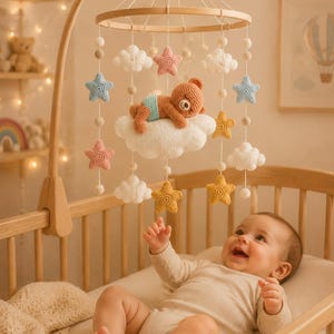 May include: A crib mobile featuring a circular wooden frame with hanging crochet elements including a brown bear sleeping on a large white cloud, surrounded by smaller white clouds and stars in shades of pink, blue, and yellow, suspended by white strings.