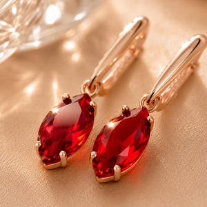 May include: A pair of rose gold drop earrings featuring marquise-cut deep red gemstones. Each earring is suspended from a polished rose gold hoop mechanism, with the faceted red stones secured by four small prongs in a classic setting design.