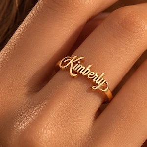 May include: A gold-toned ring featuring the name Kimberly in a cursive script font. The ring has a polished metallic finish and is worn on a finger, showcasing the elegant lettering that forms the centrepiece of the jewellery design.