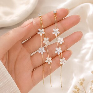 Puede incluir: Elegantes pendientes dorados con delicados detalles de flores blancas. Cada pendiente presenta una pequeña gema brillante en la parte superior y una serie de diseños florales en cascada. Los pendientes están diseñados para colgar, creando un aspecto elegante y femenino.