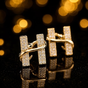 Könnte beinhalten: Ein Paar goldfarbene Manschettenknöpfe in Form des Buchstabens "H". Jeder Manschettenknopf ist mit kleinen, funkelnden Kristallen besetzt. Ein goldfarbenes, schlangenartiges Design umschlingt den Buchstaben "H". Der Hintergrund ist schwarz mit verschwommenen goldenen Lichtern.
