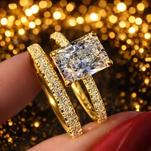 May include: A gold engagement ring set with a large, rectangular-cut diamond and a matching band. The rings are encrusted with small, sparkling stones, creating a luxurious and elegant look. The setting is a warm gold color.